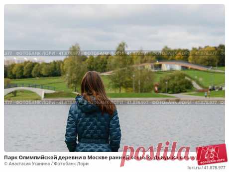 Парк Олимпийской деревни в Москве ранней осенью. Девушка в голубой куртке около пруда на фоне зелёного парка и серого неба. Стоковое фото, фотограф Анастасия Усанина / Фотобанк Лори