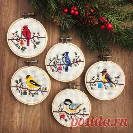 four cross stitch birds sitting on top of a wooden table next to pine cones and christmas decorations