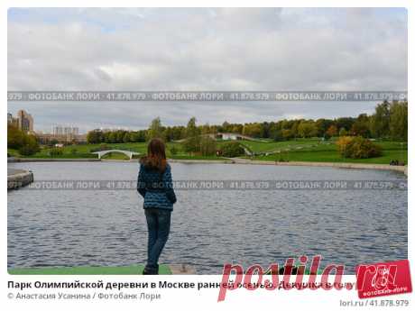 Парк Олимпийской деревни в Москве ранней осенью. Девушка в голубой куртке и синих джинсах стоит и смотрит вдаль около пруда на фоне зелёного парка и серого неба. (2016 год) Редакционное фото, фотограф Анастасия Усанина / Фотобанк Лори